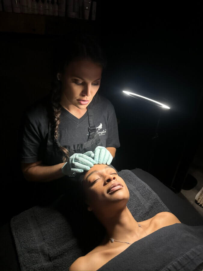 BHAM, These Are Great Last-Minute Gift Cards! Facials, Massages & More - 5 An esthetician wearing gloves performs a facial treatment on a relaxed client lying on a treatment bed under a bright lamp—an experience perfect for spa gift cards or last-minute spa gifts.