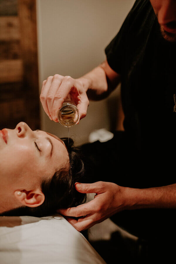 BHAM, These Are Great Last-Minute Gift Cards! Facials, Massages & More - 4 A person pours oil onto the forehead of a woman lying down with eyes closed, possibly during a spa or massage treatment—an experience you can gift with Last-Minute Spa Gift Cards.