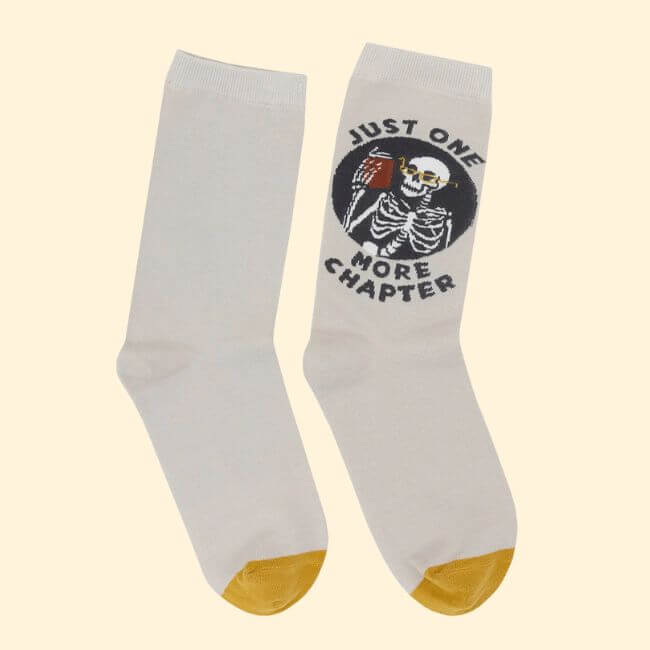 10 Unique Gift Ideas for Book Lovers - 5 A pair of white socks with yellow toes; one sock features a skeleton holding a book and the text "Just One More Chapter." Perfect gift ideas for book lovers who can't put their stories down.