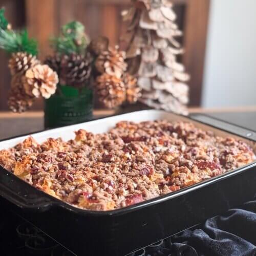 Eggnog French Toast Bake โ Perfect for the Holidays! - 6 A rectangular baking dish filled with an Overnight French Toast Bake topped with crumbly streusel, set on a dark cloth with pine cone decorations and a wooden treeโperfect for a cozy morning of Cooking Unscripted.