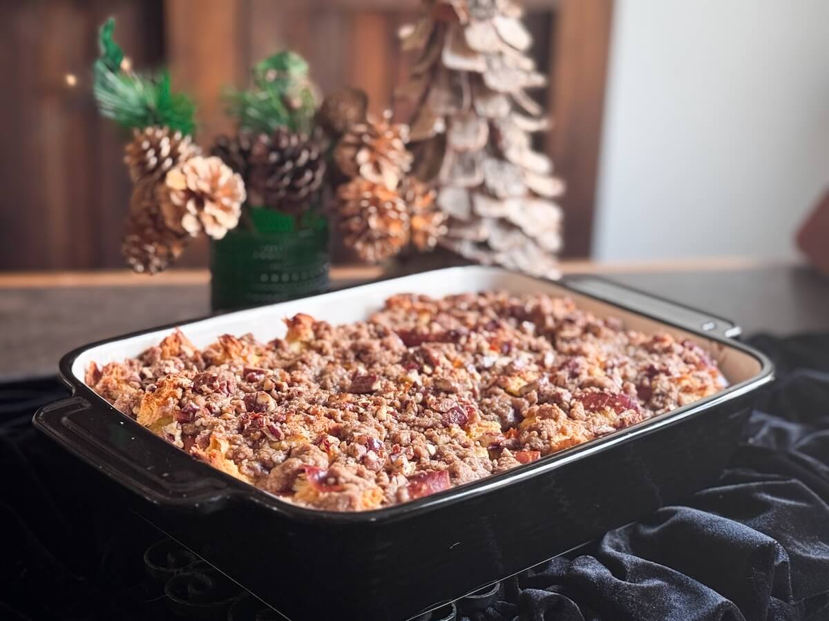 A rectangular baking dish filled with an Overnight French Toast Bake topped with crumbly streusel, set on a dark cloth with pine cone decorations and a wooden tree—perfect for a cozy morning of Cooking Unscripted.