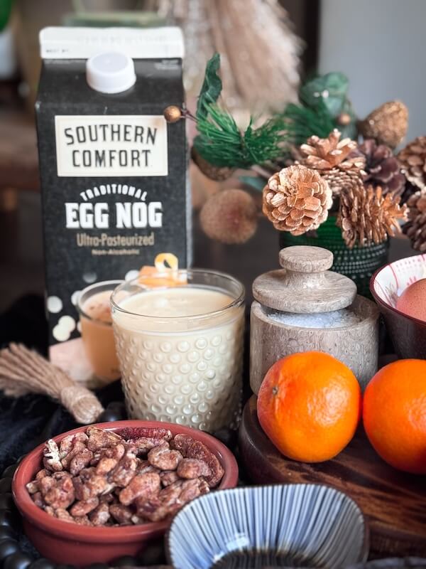 Eggnog French Toast Bake โ Perfect for the Holidays! - 3 A carton of Southern Comfort Eggnog, a glass of eggnog, two oranges, a bowl of pecans, pinecones, and decorative items arranged on a tableโperfect inspiration for your next Overnight French Toast Bake or Cooking Unscripted moment.