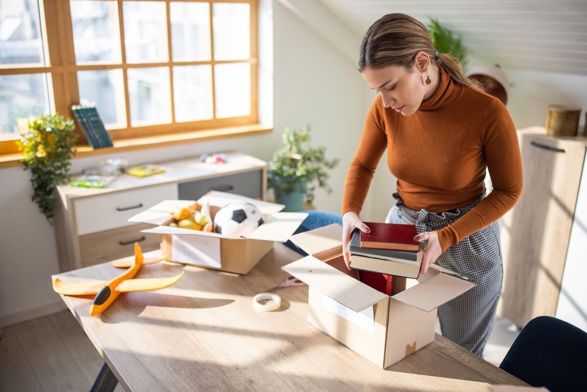 A woman in a brown turtleneck packs books and valuable items into a cardboard box on a table, with other boxes and household goods around her in a well-lit room, deciding what to sell instead of donate.
