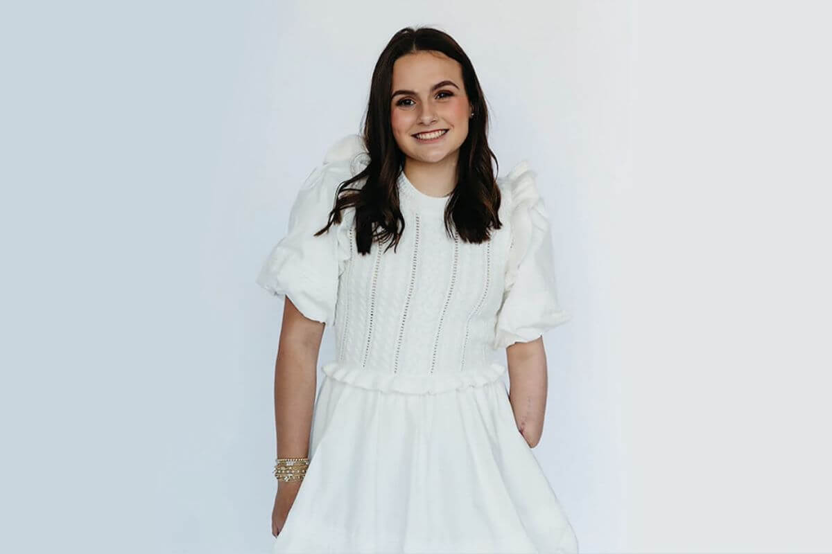 17-year-old Lulu Gribbin, a shark attack survivor with long brown hair, stands against a plain light background in a white dress with short puffed sleeves. One of her arms is partially visible.
