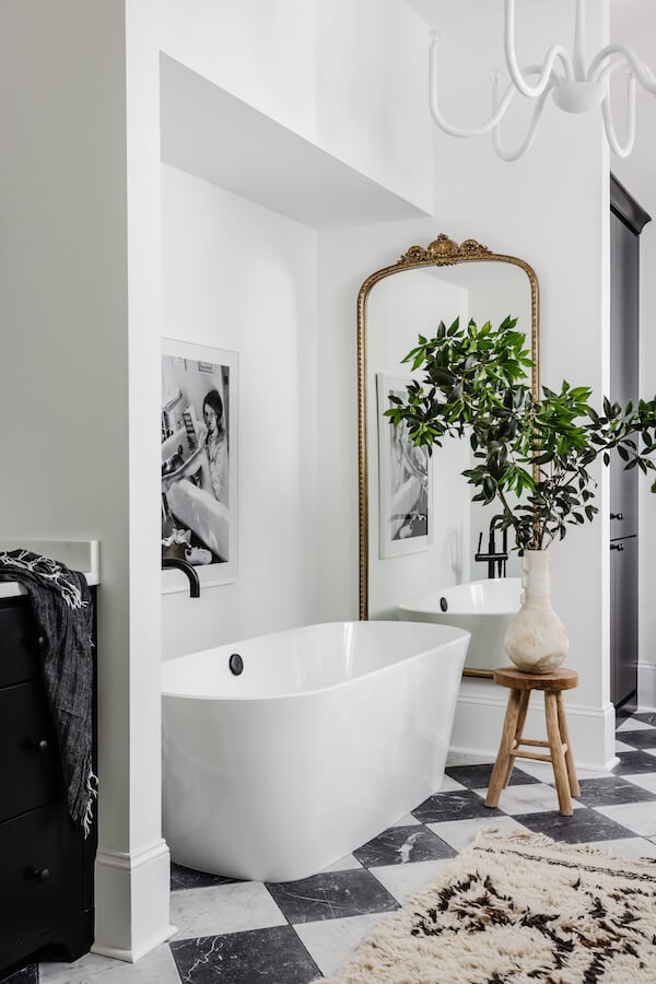 A Charlotte Home Bathed in Light, Texture, and Modern Romance - 12 Modern bathroom with a white freestanding tub, large ornate mirror, black and white photo, vase with green branches on a stool, and black-and-white checkered floor tiles.