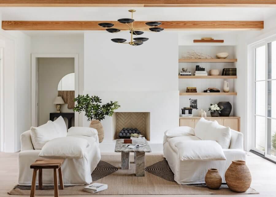 A Charlotte Home Bathed in Light, Texture, and Modern Romance - 2 Modern living room with white armchairs, a stone coffee table, built-in shelves, large vases, a fireplace, wooden ceiling beams, and a contemporary black chandelier.