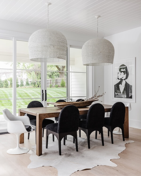 A Charlotte Home Bathed in Light, Texture, and Modern Romance - 7 Modern dining room with a wooden table, six black chairs, a white accent chair, two large pendant lights, and a monochrome portrait on the wall. Large windows overlook a green lawn.