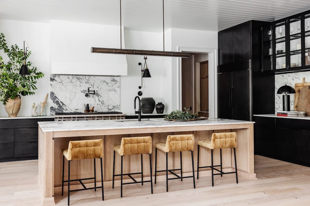 Modern kitchen with black cabinets, a large marble island, four mustard-colored barstools, and decorative plants and kitchenware on the counters.