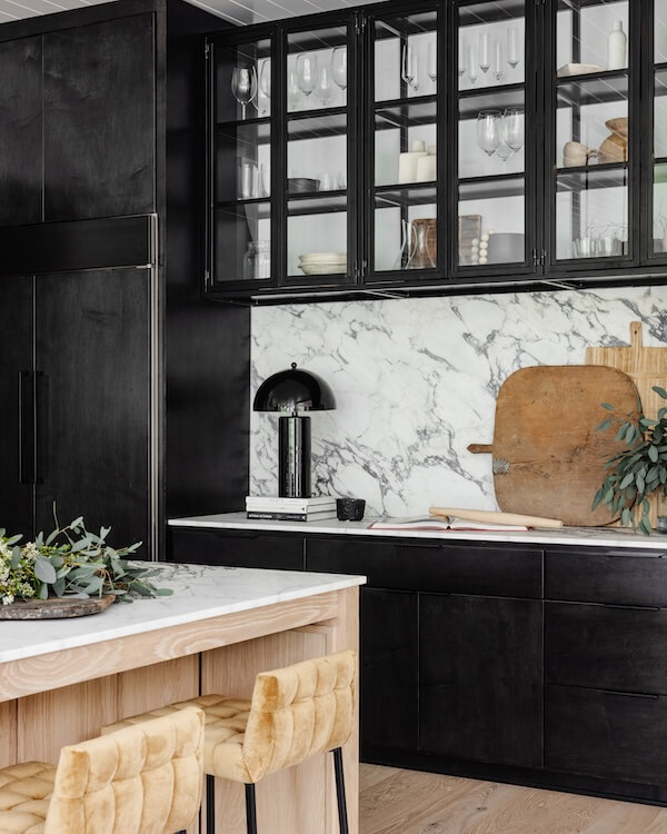 A Charlotte Home Bathed in Light, Texture, and Modern Romance - 9 Modern kitchen with black cabinets, glass upper cupboards, marble backsplash, wooden cutting boards, and a light wood island with two beige upholstered stools.