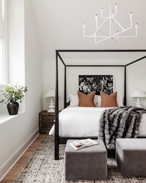 A Charlotte Home Bathed in Light, Texture, and Modern Romance - 6 Modern bedroom with a four-poster bed, brown and white bedding, faux fur throw, two gray ottomans, and a black-and-white photo above the headboard. White walls and a window with a potted plant.