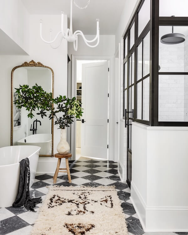 A Charlotte Home Bathed in Light, Texture, and Modern Romance - 11 A modern bathroom with black and white checkered floor, a freestanding tub, a gold-framed mirror, a vase with greenery on a stool, and a glass-enclosed shower.