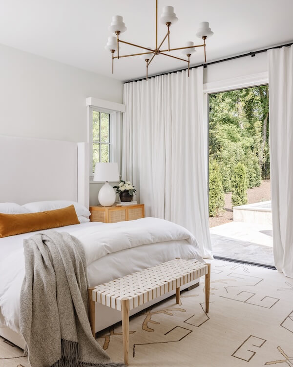 A Charlotte Home Bathed in Light, Texture, and Modern Romance - 13 Modern bedroom with white walls, bed, and curtains, a wooden bench, a brown accent pillow, bedside table with lamp, and an open door leading to a garden area.