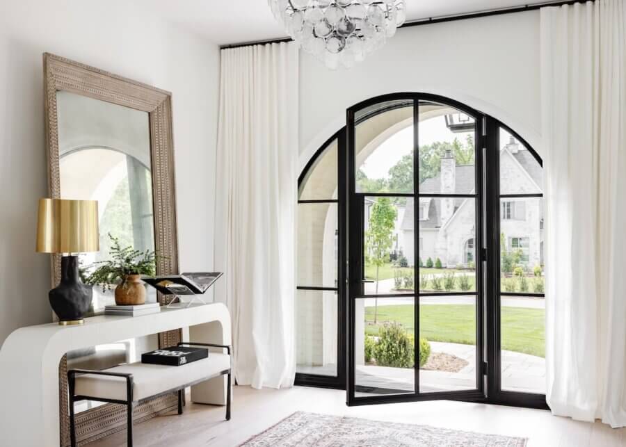 A Charlotte Home Bathed in Light, Texture, and Modern Romance - 5 Modern entryway with a large arched glass door, white walls, elegant console table, mirror, gold lamp, and view of a green lawn and neighboring house outside.