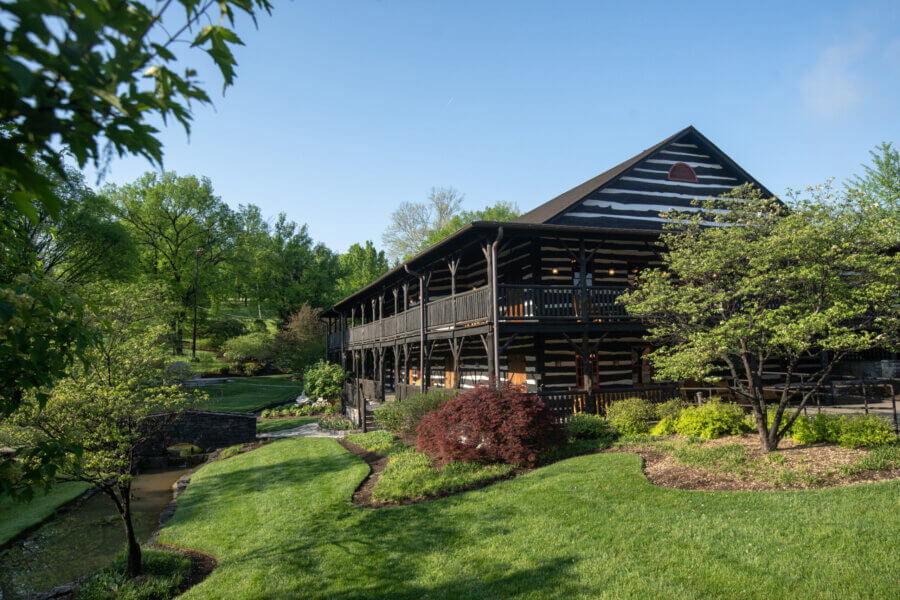 Are These Kentucky's Most Beautiful Distilleries? - 3 A large log cabin-style building with two stories sits amid manicured green lawns, landscaped shrubs, and mature trees on a clear day.