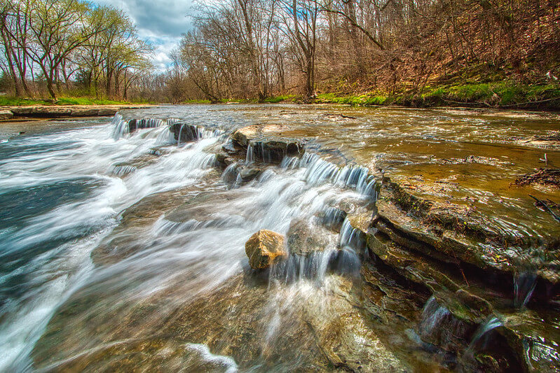 Are These Kentucky's Most Beautiful Distilleries? - 12 A shallow stream flows over layered rocks, creating small waterfalls, with bare trees and early spring greenery along the banks under a partly cloudy sky.