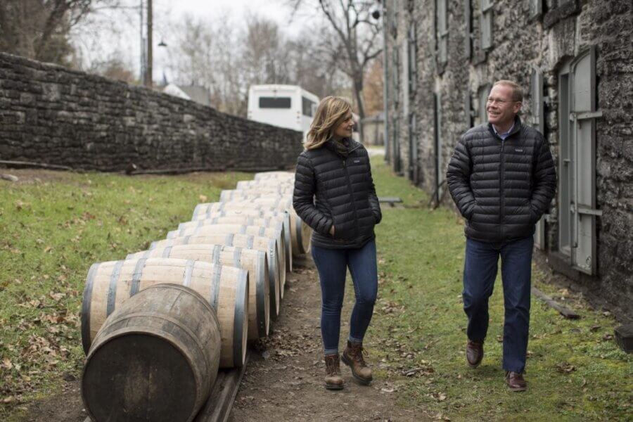 Are These Kentucky's Most Beautiful Distilleries? - 14 Two people walk and talk beside a row of wooden barrels outside a stone building on a grassy path, capturing the charm of Kentucky distilleries.