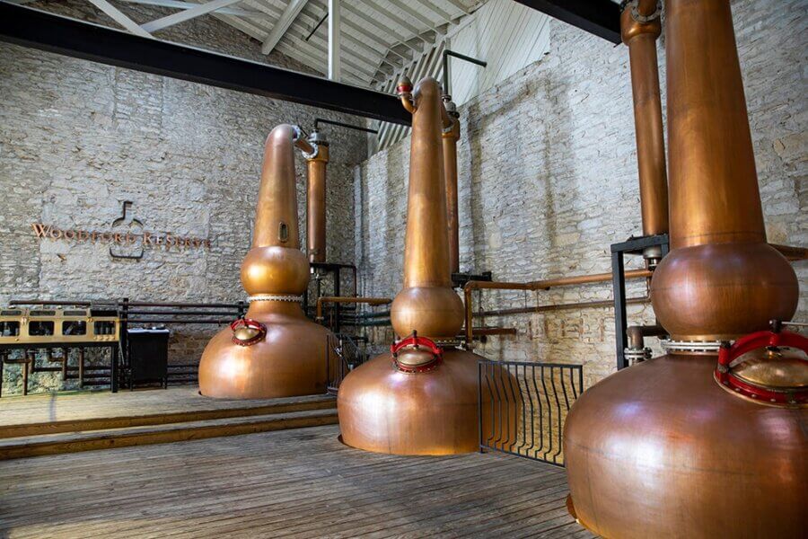 Are These Kentucky's Most Beautiful Distilleries? - 15 Three large copper stills stand in a distillery with stone walls, wooden floors, and the Woodford Reserve logo on the back wall.