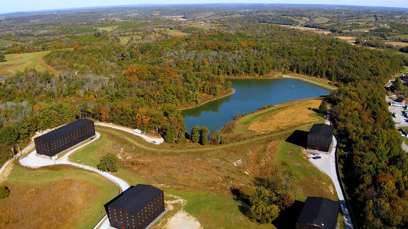 Are These Kentucky's Most Beautiful Distilleries? - 11 Aerial view of a rural landscape featuring a small lake surrounded by trees, open fields, and several rectangular buildings along winding roads.
