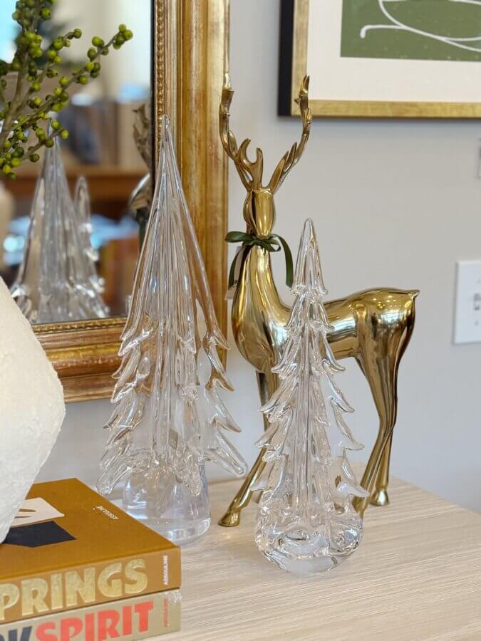 23 Luxurious Nashville FINDS for Holiday 2025 - 16 A gold reindeer figurine and two glass Christmas tree decorations are displayed on a wooden surface next to stacked books and framed artwork, offering inspiration for elegant holiday decor and expensive gift ideas.