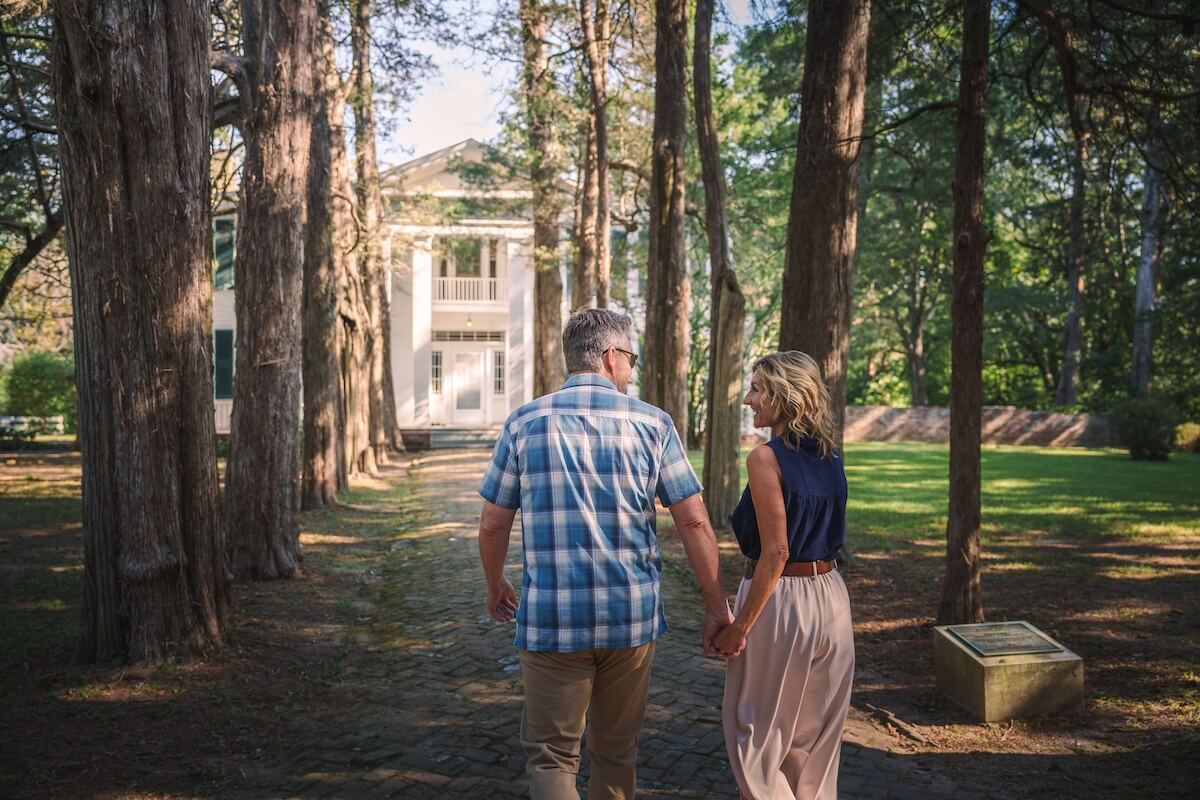 An older couple walks hand in hand down a tree-lined path toward a large white house on a sunny day, creating memorable moments as they explore charming Mississippi destinations together.
