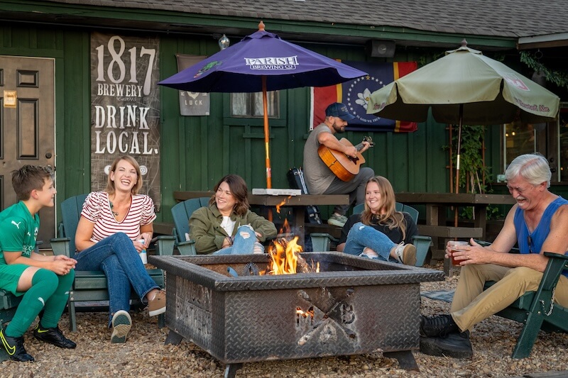8 Mississippi Destinations Worth the Drive - 14 A group of people sits around a fire pit outside a green building, talking and laughing as a person plays guitar in the backgroundβcreating memorable moments at one of the unique Mississippi destinations for travel.