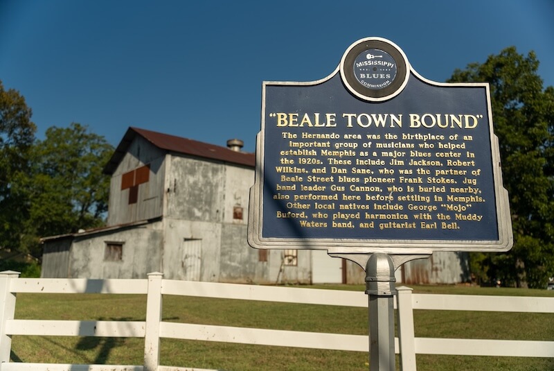 8 Mississippi Destinations Worth the Drive - 10 A historical marker titled "Beale Town Bound" stands in front of a white fence and weathered barn, inviting travelers to explore one of the memorable moments in Mississippi destinations and discover the area's rich musical heritage.