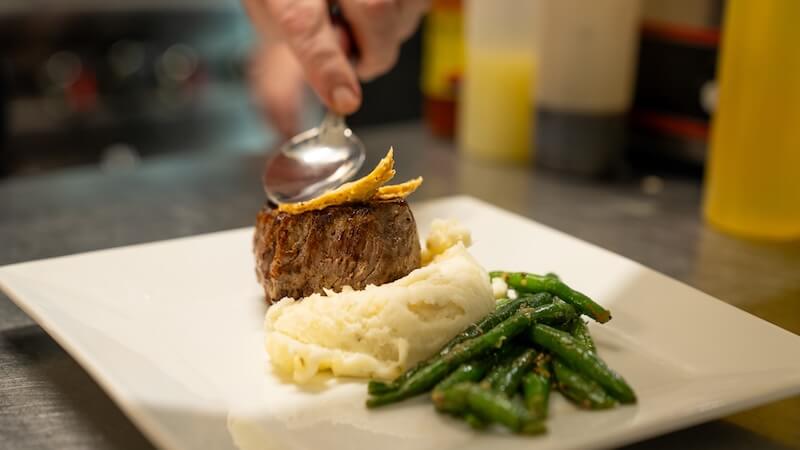 8 Mississippi Destinations Worth the Drive - 11 A plated meal featuring a portion of steak on mashed potatoes, green beans on the side, and a hand placing a garnish on topβperfect for savoring memorable moments during your Mississippi travel adventures.