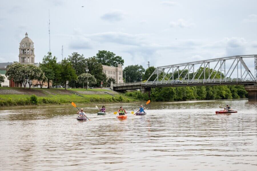 8 Mississippi Destinations Worth the Drive - 12 Five people kayak on a river near a metal bridge and a town with a clock tower, under a cloudy skyβcapturing the memorable moments that make Mississippi destinations perfect for travel.