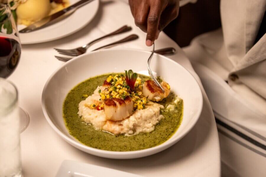 8 Mississippi Destinations Worth the Drive - 8 A person enjoys a forkful of seared scallops atop risotto, garnished with corn and herbs, surrounded by green sauceβa memorable moment perfect for those seeking unique Mississippi destinations.