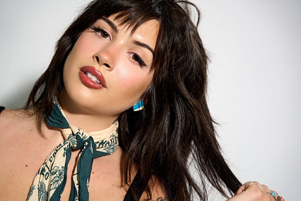 Mara Daniele, featured in FACES, gazes at the camera with dark hair and a patterned scarf around her neck, holding a section of her hair with one hand against a plain background.