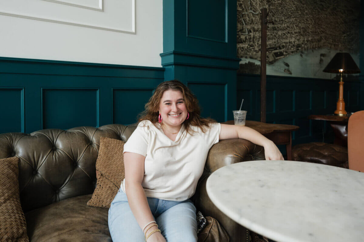 Cecilia Fay sits on a leather couch in a modern, stylish room, smiling at the camera. A drink and a round white table are in the foreground, capturing one of the many expressive FACES of contemporary lifestyle.