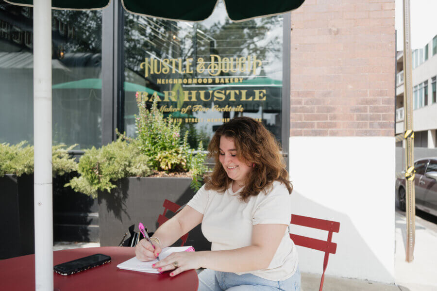 Meet Cecilia Fay of Women Connect Memphis - 2 Cecilia Fay sits at a red outdoor table, writing in a notebook with a pink pen. A phone and notebook are on the table. The FACES of Hustle & Dough bakery's storefront appear in the background.