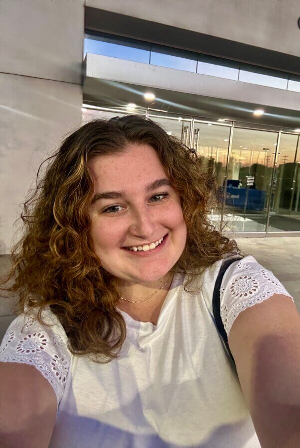 Meet Cecilia Fay of Women Connect Memphis - 5 Cecilia Fay, part of FACES, smiles at the camera with her curly hair and nose ring, standing outside a modern glass building in the evening.