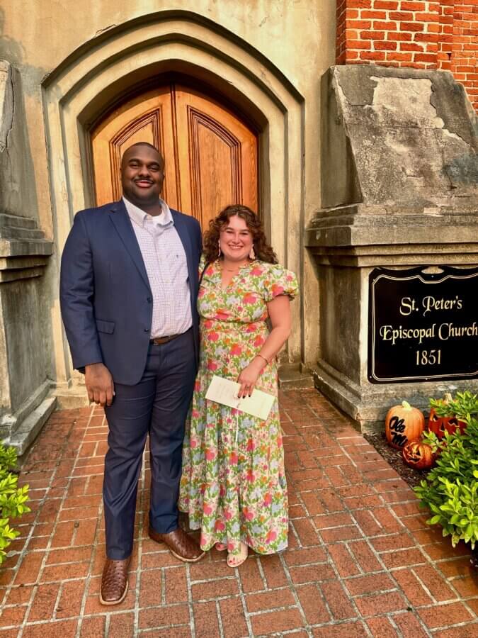Meet Cecilia Fay of Women Connect Memphis - 7 A man in a suit and Cecilia Fay in a floral dress stand smiling in front of St. Peter’s Episcopal Church, 1851, next to a sign and fall decorations—capturing joyful FACES on a crisp autumn day.