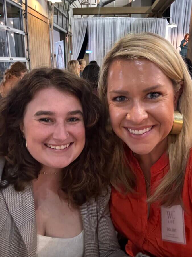Meet Cecilia Fay of Women Connect Memphis - 4 Cecilia Fay and another woman smile at the camera while seated indoors at a FACES event, with other attendees and curtains visible in the background.