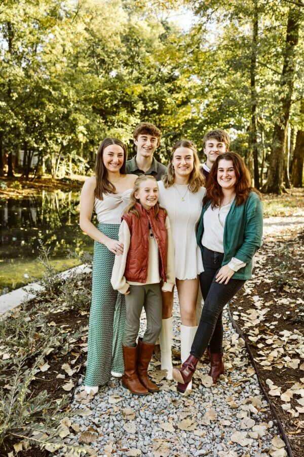 Meet Cecilia Fay of Women Connect Memphis - 3 Six people stand together smiling outdoors on a gravel path beside a pond, surrounded by trees and fallen leaves in a wooded area, enjoying the moment with FACES full of joy.