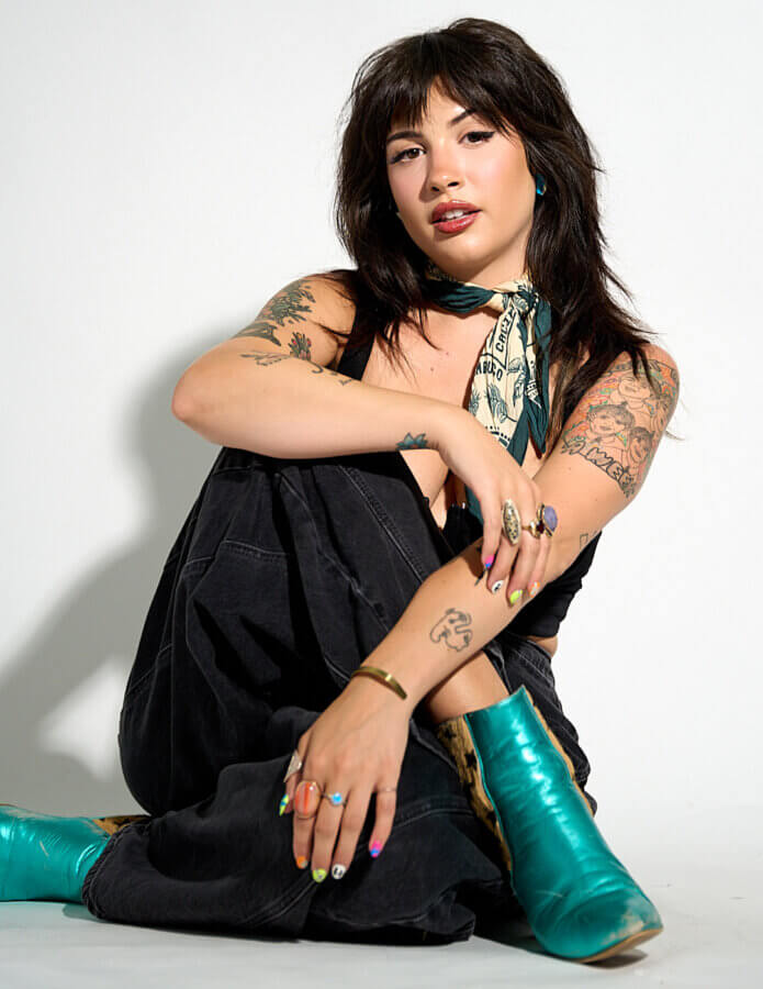 She Sees Magic in Every Corner of Memphis - 2 Woman with dark hair and tattoos sits on the floor against a white background, wearing teal boots, dark pants, a scarf, and colorful rings and nail polish.