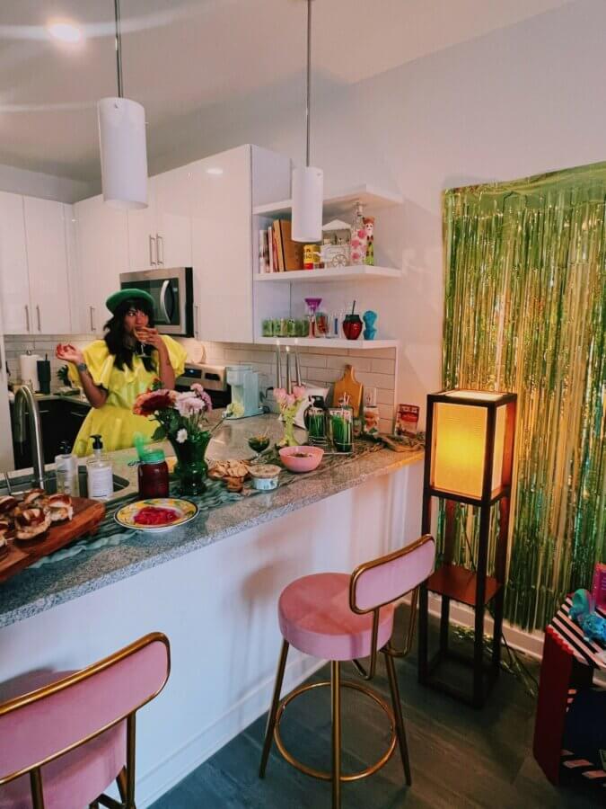 She Sees Magic in Every Corner of Memphis - 3 A woman in a bright yellow dress and green hat stands in a modern kitchen decorated with snacks, flowers, and festive green tinsel on the wall. Pink barstools are at the counter.