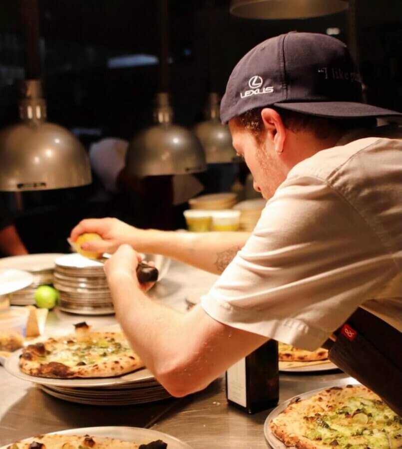 Memphis’s 10 Most Popular Articles of 2025 - 5 A chef wearing a cap prepares pizzas at a kitchen counter in Memphis, adding toppings under hanging lights, with plates and ingredients in the background.