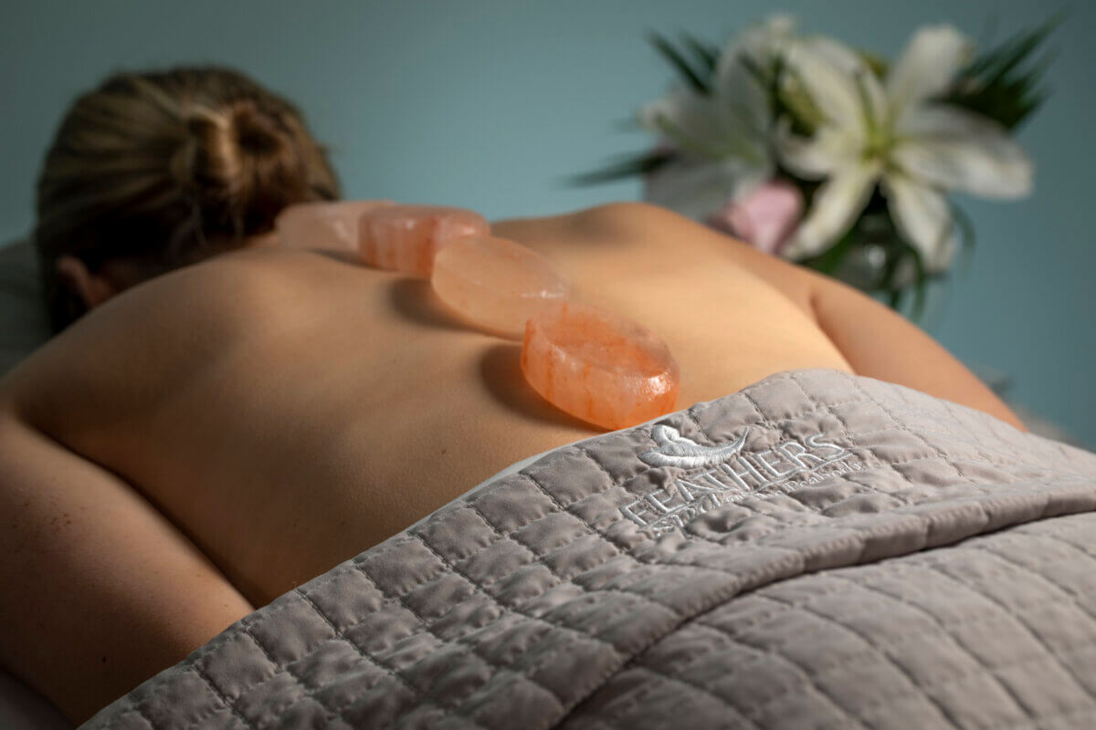 Relax at one of the best day spas in Memphis: a person lies face down on a massage table, three large salt stones on their back, surrounded by pampering touches like a floral arrangement and "FEATHERS" embroidered on the blanket.