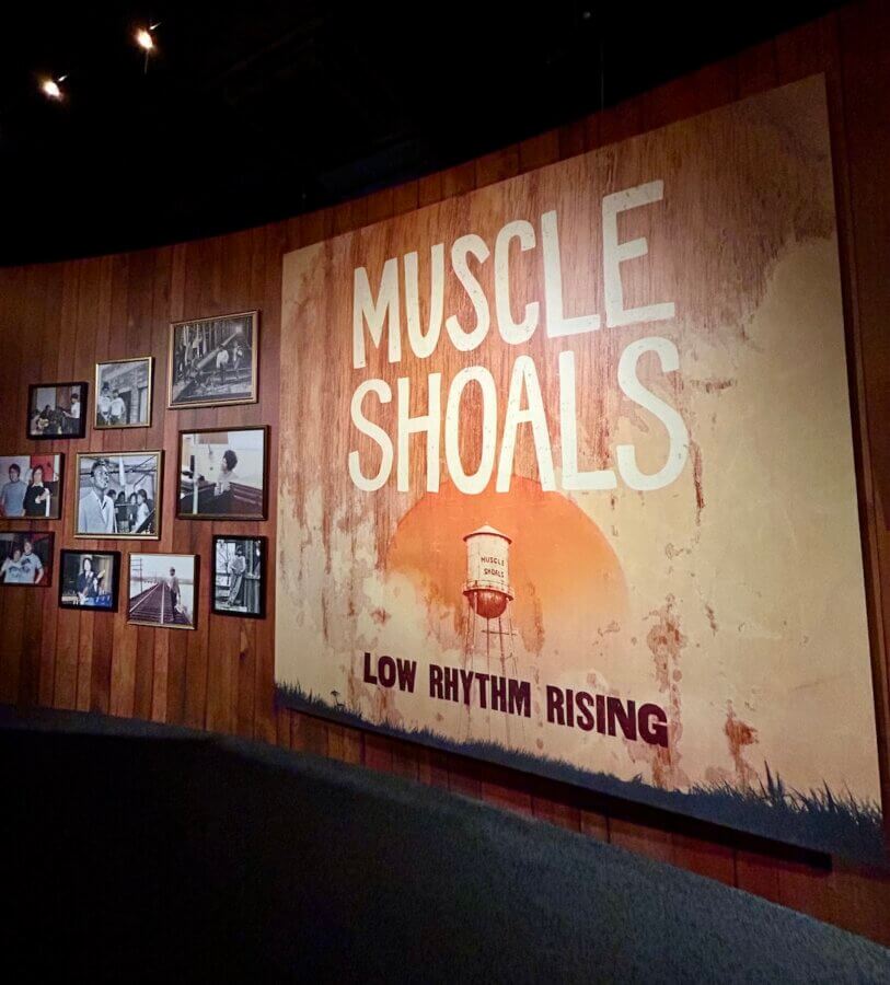 The Country Music Hall of Fame Unveils Its NEW Muscle Shoals Exhibit - 2 Large "MUSCLE SHOALS LOW RHYTHM RISING" mural on a curved wall, with framed historical photos displayed on adjacent wooden panels—part of the new Muscle Shoals exhibit at the Country Music Hall of Fame.