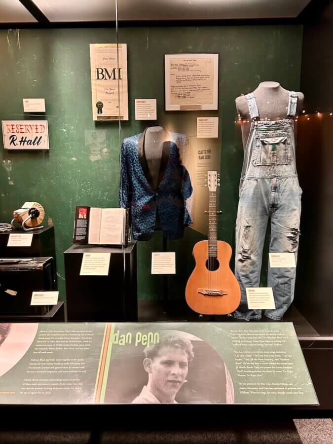 The Country Music Hall of Fame Unveils Its NEW Muscle Shoals Exhibit - 3 Exhibit at the Country Music Hall of Fame showcasing a blue patterned jacket, denim overalls, an acoustic guitar, framed documents, and memorabilia celebrating Muscle Shoals musician Dan Penn.
