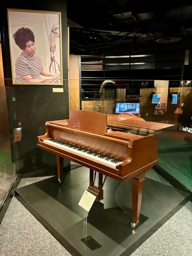 The Country Music Hall of Fame Unveils Its NEW Muscle Shoals Exhibit - 4 A brown Yamaha piano is displayed in the Country Music Hall of Fame’s Muscle Shoals Exhibit, with a framed photo of a woman singing into a microphone mounted on the wall above it, highlighting country music’s rich heritage.