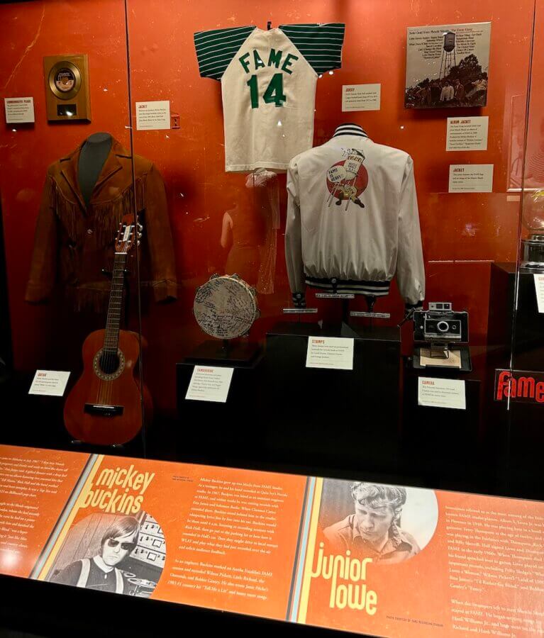 The Country Music Hall of Fame Unveils Its NEW Muscle Shoals Exhibit - 5 A new exhibit at the Country Music Hall of Fame features a museum display case with a guitar, fringe jacket, tambourine, Fame 14 jersey, and more, highlighting Mickey Buckins and Junior Lowe in this Muscle Shoals exhibit.