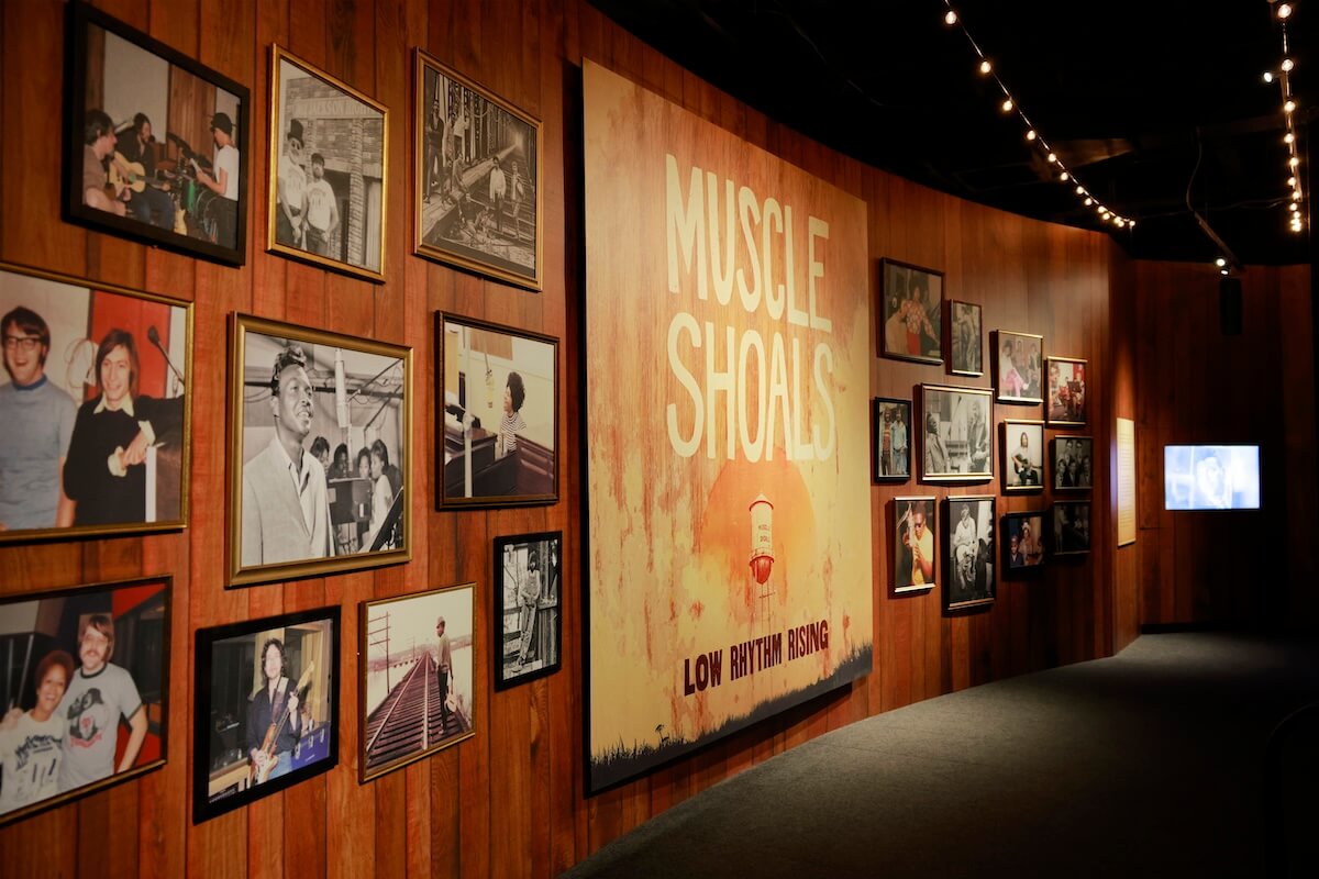 A museum exhibit wall at the Country Music Hall of Fame features photos and a large sign reading "Muscle Shoals: Low Rhythm Rising," with string lights above and framed pictures of musicians and recording sessions.