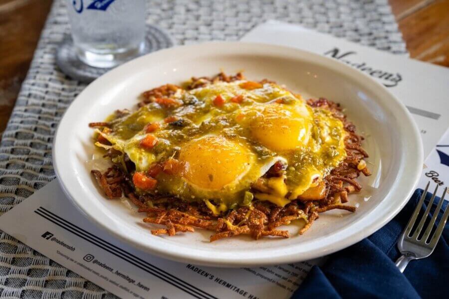 A plate of hash browns topped with two sunny-side-up eggs, green chili sauce, diced tomatoes, and melted cheese, served on a woven placemat next to a glass of water—a true Hermitage dining favorite recommended in any local's guide.