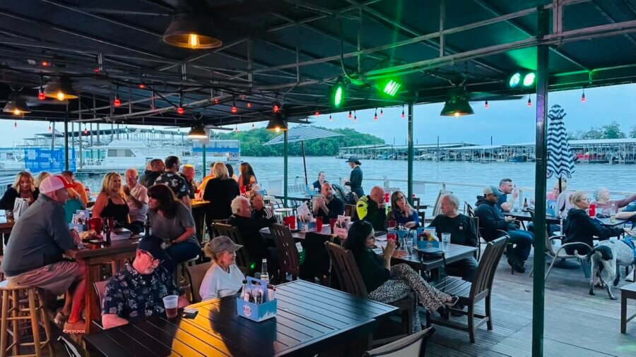 People sit at tables and socialize under a covered patio overlooking a lake, with boats and docks visible in the background, enjoying the relaxed atmosphere typical of Hermitage dining.