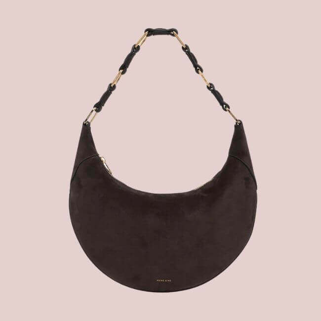 23 Luxurious Nashville FINDS for Holiday 2025 - 3 A dark brown crescent-shaped suede handbag with a short handle featuring gold and black chain links, displayed on a light beige background—perfect for those seeking expensive gift ideas.