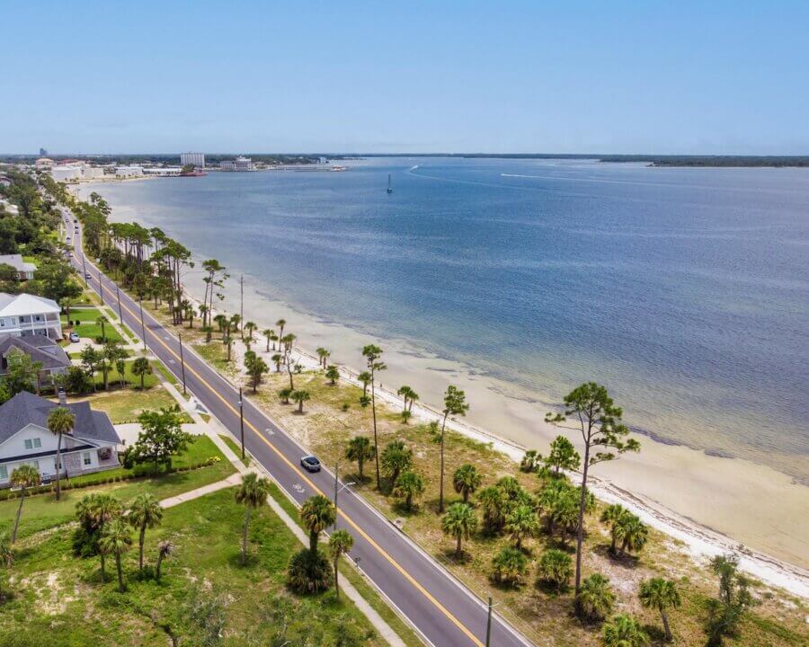 Panama City’s Multi-Coastal Story: History, Culture & Life on the Bay - 2 A coastal road in Panama City FL runs parallel to a sandy shoreline with palm trees, houses on the left, and calm blue water on the right under a clear sky—a perfect spot for travel and tourism.