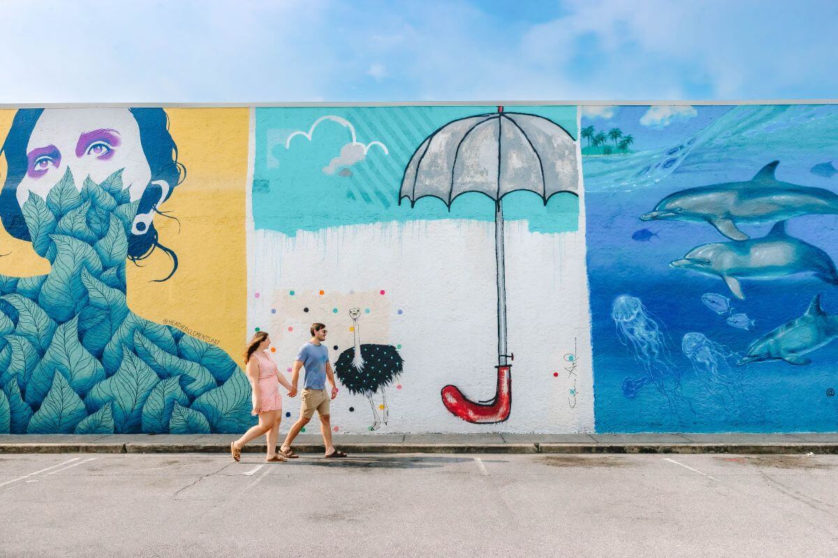 Two people walk past a colorful mural in Panama City FL, featuring a woman with leaves, an umbrella with a red handle, and dolphins swimming underwater—capturing the vibrant spirit of Panama City tourism.